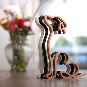 K&ouml;nnte beinhalten: Eine mehrschichtige Holzskulptur eines Hundes in Wei&szlig;, Schwarz, Beige und Grau. Der Hund ist stilisiert mit einer geschwungenen Silhouette, und die Schichten erzeugen einen 3D-Effekt. Die Skulptur steht auf einer Holzoberfl&auml;che, mit einem verschwommenen Hintergrund von Blumen.