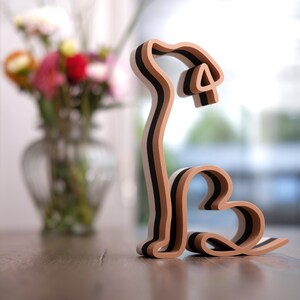 K&ouml;nnte beinhalten: Holzhund-Skulptur mit herzf&ouml;rmigem K&ouml;rper und gebogenem Schwanz. Die Skulptur besteht aus abwechselnden Schichten aus hellbraunem und schwarzem Holz. Der Hund sitzt auf einer Holzoberfl&auml;che, im Hintergrund eine Vase mit Blumen.