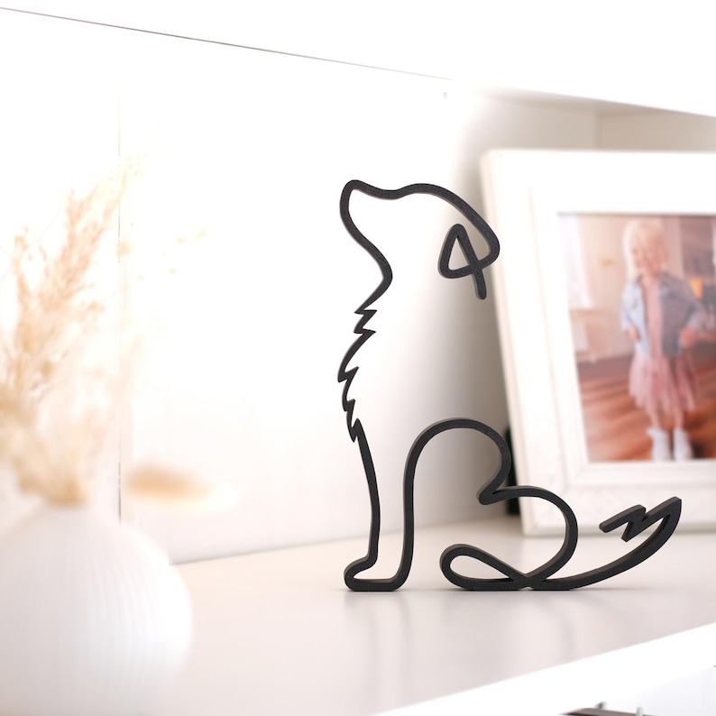 K&ouml;nnte beinhalten: Schwarze Metallskulptur eines Hundes im Sitzen, mit erhobenem Kopf und herzf&ouml;rmigem Schwanz. Die Skulptur steht auf einer wei&szlig;en Oberfl&auml;che, im Hintergrund eine wei&szlig;e Vase und ein gerahmtes Foto.