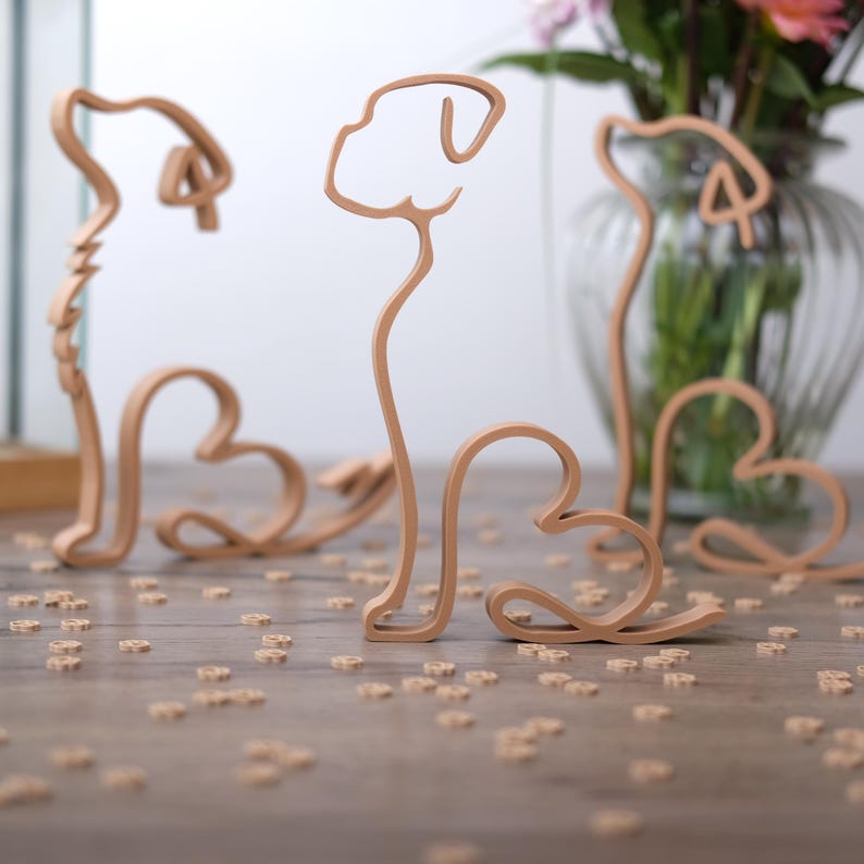 K&ouml;nnte beinhalten: Drei hellbraune Holzskulpturen von Hunden mit herzf&ouml;rmigen K&ouml;rpern und einfachen Linien. Sie stehen auf einer Holzoberfl&auml;che, die mit kleinen, herzf&ouml;rmigen Ausschnitten &uuml;bers&auml;t ist. Im Hintergrund befindet sich eine Glasvase mit Blumen.