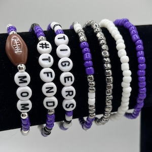 May include: A set of six beaded bracelets featuring purple, white, and metallic silver beads. One bracelet includes a brown football charm and the word MOM. Others feature white letter beads spelling TIGERS and #TFND, alongside solid purple, white, and silver strands.