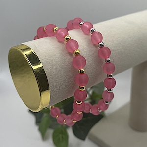 May include: Two pink beaded bracelets with gold and silver accents. The bracelets are displayed on a white stand with a gold base. The beads are a matte finish and appear to be made of glass or a similar material.