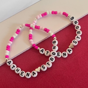 May include: Two beaded bracelets with white letter beads spelling "ATHENA STRONG" and "ATOENA STRONG". The bracelets feature pink, white, and silver beads. Each bracelet has a small gold heart charm. The bracelets are on a red surface.