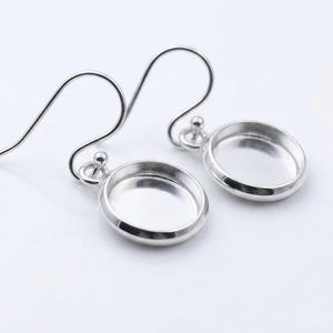 Könnte beinhalten: Ein Paar silberfarbene Ohrringe mit Haken-Design. Jeder Ohrring hat eine kleine, runde, vertiefte Fläche, ideal zum Hinzufügen dekorativer Elemente. Die Ohrringe werden vor einem schlichten weißen Hintergrund gezeigt.