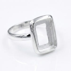 May include: A silver ring with a rectangular bezel setting. The ring has a polished finish and a simple band. The setting is designed to hold a rectangular gemstone or other decorative element.