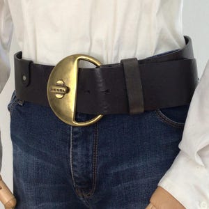Peut inclure: Une ceinture en cuir marron foncé dotée d'une grande boucle en laiton semi-circulaire avec un mécanisme de verrouillage rotatif central. La boucle porte l'inscription DIESEL. La ceinture est portée sur un jean bleu et une chemise blanche.