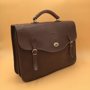 May include: A brown leather briefcase with a top handle and silver-tone hardware. The bag features two buckle closures on the front flap and a small logo. The briefcase is designed with a classic, professional aesthetic.