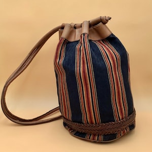 May include: A cylindrical drawstring bucket bag featuring a vertical striped pattern in navy blue, red, and cream. The bag includes brown leather trim at the top opening, a braided leather band around the base, and long brown leather shoulder straps.
