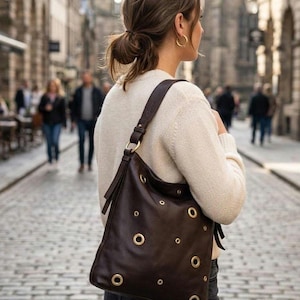 Può includere: Borsa a tracolla in pelle marrone scuro con occhielli color oro. La borsa ha una lunga tracolla e si indossa a spalla. Una persona porta la borsa mentre cammina per una strada acciottolata.