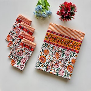 Puede incluir: Un juego de cinco servilletas de algodón blanco y un mantel a juego. Las servilletas y el mantel presentan un estampado floral en naranja, rojo y verde. El borde superior de las servilletas y el mantel es de rayas naranjas y blancas.