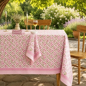 Puede incluir: Un mantel rectangular con un patrón floral rosa repetitivo sobre fondo blanco con un diseño de celosía verde. La tela tiene un borde ancho de color rosa con rayas finas. Una pila de servilletas a juego descansa sobre la mesa.