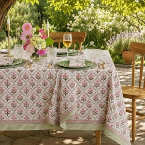 Può includere: Una tovaglia rettangolare con un motivo a rombi ripetuto con disegni floreali rosa e steli verdi. Il tessuto presenta un ampio bordo a righe verdi lungo i lati. La tavola è apparecchiata con piatti verdi, bicchieri trasparenti e un centrotavola.