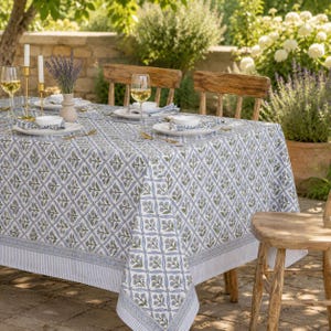 May include: A rectangular tablecloth featuring a repeating blue and green floral block print pattern on a white background with a striped border. The table is set with white plates, clear wine glasses, and a vase of dried lavender flowers.