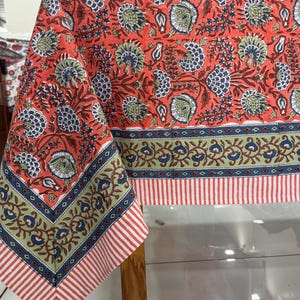 May include: A coral-colored tablecloth with a floral pattern in blue, green, and white. The border features a striped design in red and white, with a blue and green floral band. The tablecloth is draped over a wooden table.