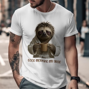 May include: White t-shirt with a sloth illustration in a bathrobe, holding a mug. The text "GOOD MORNING MY ARSE" is printed below the sloth image. The sloth has a tired expression. The shirt is worn by a person.