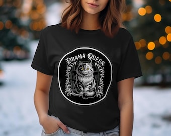 Könnte beinhalten: Schwarzes T-Shirt mit einem kreisförmigen Aufdruck. Der Aufdruck zeigt eine detaillierte Illustration einer Katze mit grünen Augen und den Worten "Drama Queen". Die Katze ist schwarz und weiß. Das Design befindet sich vor einem verschwommenen Hintergrund.