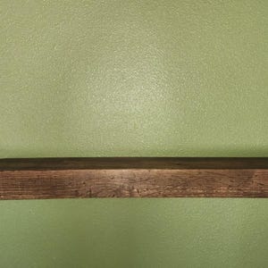 May include: A rustic, rectangular wooden shelf, stained a dark brown, is mounted against a textured, olive green wall. The shelf has a simple, minimalist design, perfect for displaying items. The shelf is approximately 30 inches long.