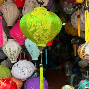 Può includere: Una lanterna verde brillante con intricati motivi floreali e geometrici, appesa tra una collezione di lanterne di carta colorate. La lanterna ha una lunga nappa giallo brillante. Altre lanterne sono rosse, rosa, bianche e gialle.