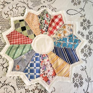 May include: A handmade, star-shaped quilted trivet with a white center and scalloped edges. The trivet is composed of various fabric patterns, including gingham, floral, and striped designs in red, green, blue, and yellow.