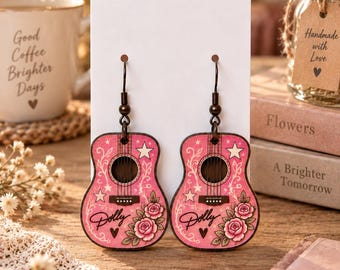 May include: A pair of dangle earrings shaped like acoustic guitars with a pink finish. The design features white stars, swirling vine patterns, and pink rose illustrations. The word Dolly is printed in black script above a small black heart on each.