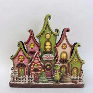 Könnte beinhalten: Eine dekorative Tischdekoration mit einer Whoville-Dorfkulisse und bunten Häusern in Grün, Rosa und Creme. Der Grinch steht davor, mit Schildern, auf denen Welcome to Whoville, Be Merry Be Grinchy, Merry Grinchmas und Grinch Approved steht.