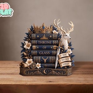 Könnte beinhalten: Ein dekoratives Display mit einem Stapel aus fünf dunkelblauen Büchern mit dem Titel Throne of Glass, gekrönt von einer goldenen Krone. Ein weißer Hirschkopf, ein silbernes Schwert, weiße Blumen und eine Schriftrolle mit dem Text YOU DO NOT YIELD.