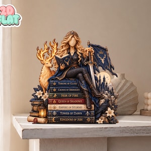 Könnte beinhalten: Eine detaillierte Harzfigur, die eine langhaarige Gestalt zeigt, welche auf einem Stapel von sieben Büchern mit den Titeln Throne of Glass, Crown of Midnight, Heir of Fire, Queen of Shadows, Empire of Storms, Tower of Dawn und Kingdom of Ash sitzt.