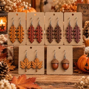 May include: Six pairs of dangle earrings displayed on brown cardboard cards. The collection includes four pairs of oak leaf shapes in yellow, orange, red, and brown, one pair of maple leaf shapes in orange, and one pair of brown acorn shapes.