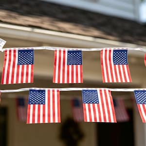 Könnte beinhalten: Eine Girlande aus kleinen amerikanischen Flaggen, jede mit einem blauen Kanton und rot-weißen Streifen, auf einer weißen Schnur aufgereiht. Die Flaggen werden im Freien ausgestellt, wahrscheinlich für eine patriotische Feier.