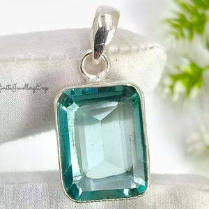 Pode incluir: Pingente retangular com uma gema de cor verde-azulado em uma moldura de prata. A pedra tem uma superfície facetada. A argola de prata é fixada na parte superior. O texto "Infinita Jewellery Exp" é visível. Joalheria.