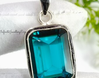 May include: A rectangular pendant with a large, teal-colored gemstone set in a silver-toned metal frame. The pendant has a bail at the top for a chain. The background is a white fabric with green foliage.