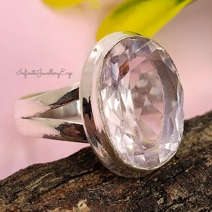 Puede incluir: Un anillo de plata con una gema grande, ovalada, facetada y transparente. El anillo tiene un diseño de caña dividida. El fondo es una mezcla de rosa, verde y marrón. El texto "Infinite Jewellery Exp" es visible.