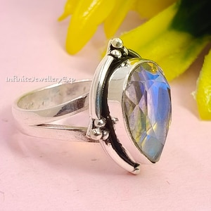Puede incluir: Un anillo de plata con una piedra preciosa iridiscente en forma de pera. El anillo tiene una caña dividida y detalles decorativos cerca del engaste de la piedra. El fondo es rosa claro con elementos florales amarillos y verdes. El texto "InfiniteJewelleryExp" es visible.