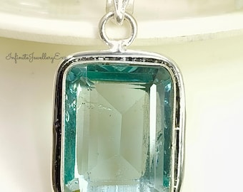 May include: A rectangular pendant with a large, light teal gemstone set in a silver-colored metal frame. The gemstone has a faceted surface. The pendant has a loop at the top for a chain. The text "Infinite Jewellery Ex" is visible.