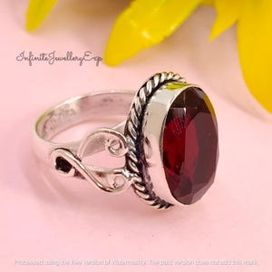 May include: A silver ring featuring a large, oval-cut, deep red gemstone. The ring has a decorative, twisted rope design around the gemstone and ornate swirls on the band. The text "Infinite Jewellery Exp" is visible.