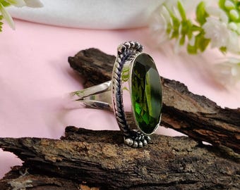 May include: A sterling silver ring featuring a large, oval-cut, deep green gemstone. The ring has a detailed, textured band with a decorative design around the gemstone. The ring is displayed on a piece of dark wood against a pink background.