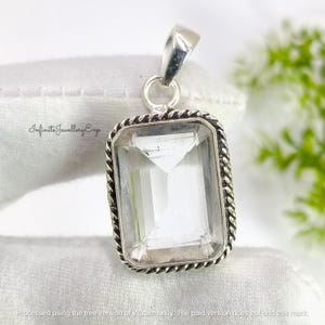 May include: A clear, rectangular gemstone pendant with a silver-coloured frame. The frame features a twisted rope design. The pendant has a loop for a chain. The text "Infinite Jewellery Exp" is visible. The pendant is approximately 2.5 cm in length.