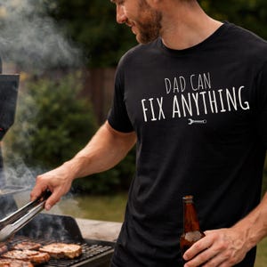Puede incluir: Camiseta negra con el texto blanco "DAD CAN FIX ANYTHING" y un pequeño gráfico de llave inglesa. La camiseta se usa mientras se hace una barbacoa al aire libre. Se sostiene una botella de cerveza en la mano.