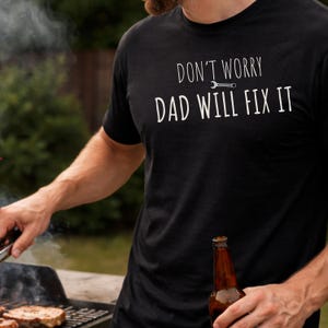 Könnte beinhalten: Schwarzes T-Shirt mit dem weißen Text "DON'T WORRY DAD WILL FIX IT". Das Bild zeigt eine Person, die im Freien grillt und eine Bierflasche und eine Zange hält.