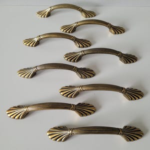 May include: A set of seven antique brass-toned metal drawer pulls. Each handle features a curved, ribbed centre bar and flared, fan-shaped ends with detailed ridges. The hardware has a weathered, metallic finish with dark highlights in the grooves.