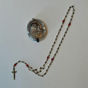 May include: A rosary necklace featuring clear glass beads interspersed with small red beads, ending in a metal crucifix. Beside it lies an oval-shaped metal locket with a raised relief depiction of a figure wearing a head covering on its front.