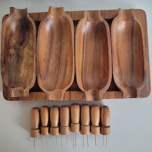 Puede incluir: Una bandeja de madera con cuatro compartimentos alargados en forma de cuenco. Debajo de la bandeja hay ocho marcadores de queso de madera con púas metálicas. La madera tiene un tono cálido y natural.