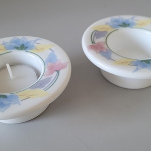 May include: Two white ceramic tea light candle holders with a flat, circular rim featuring a painted floral pattern in shades of blue, yellow, and pink. One holder contains a small, round white tea light candle with a visible white wick.