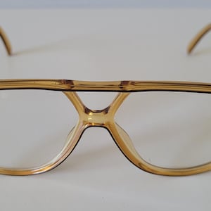 May include: A pair of amber-coloured aviator-style spectacles with a translucent plastic frame. The glasses feature a prominent double bridge design across the top of the lenses and thin, curved temples extending from the sides of the frame.