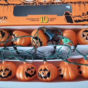May include: A set of 10 orange plastic pumpkin-shaped string lights in a cardboard box labelled Danson Decor. The pumpkins feature black cut-out jack-o'-lantern faces and are connected by a green electrical cord with a standard two-prong plug.