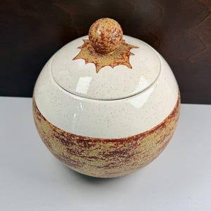 May include: A spherical ceramic jar with a lid featuring a round knob. The upper portion is a speckled cream colour, while the lower section and the area around the knob display a textured, mottled reddish-brown and tan glaze pattern.