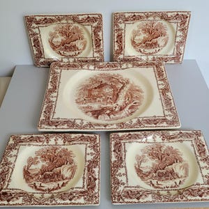May include: A collection of five antique cream-coloured plates with a brown transferware design. The plates display a central landscape scene and a decorative border. The set includes four square plates and one rectangular serving dish.