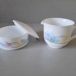 May include: A white ceramic sugar bowl with a matching lid and a creamer jug. Both pieces feature a delicate floral pattern with blue, pink, and yellow leaves and petals. The creamer has a curved handle and a pouring spout, while the sugar bowl is round.