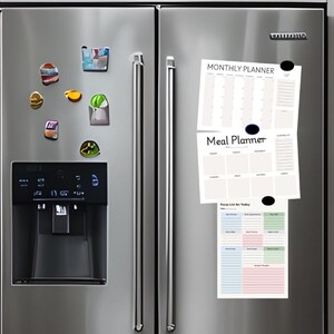 Op de afbeelding: Een roestvrijstalen koelkastdeur met diverse kleurrijke magneten. Er zijn drie witte papieren planners bevestigd: een maandplanner, een maaltijdplanner en een focuslijst voor vandaag.