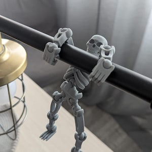 Könnte beinhalten: Eine graue, bewegliche Skelettfigur, die sich an einer schwarzen Stange festhält. Das Skelett hat einen detaillierten Schädel und ist so positioniert, als würde es hängen. Die Figur ist aus Kunststoff und ein dekorativer Gegenstand.